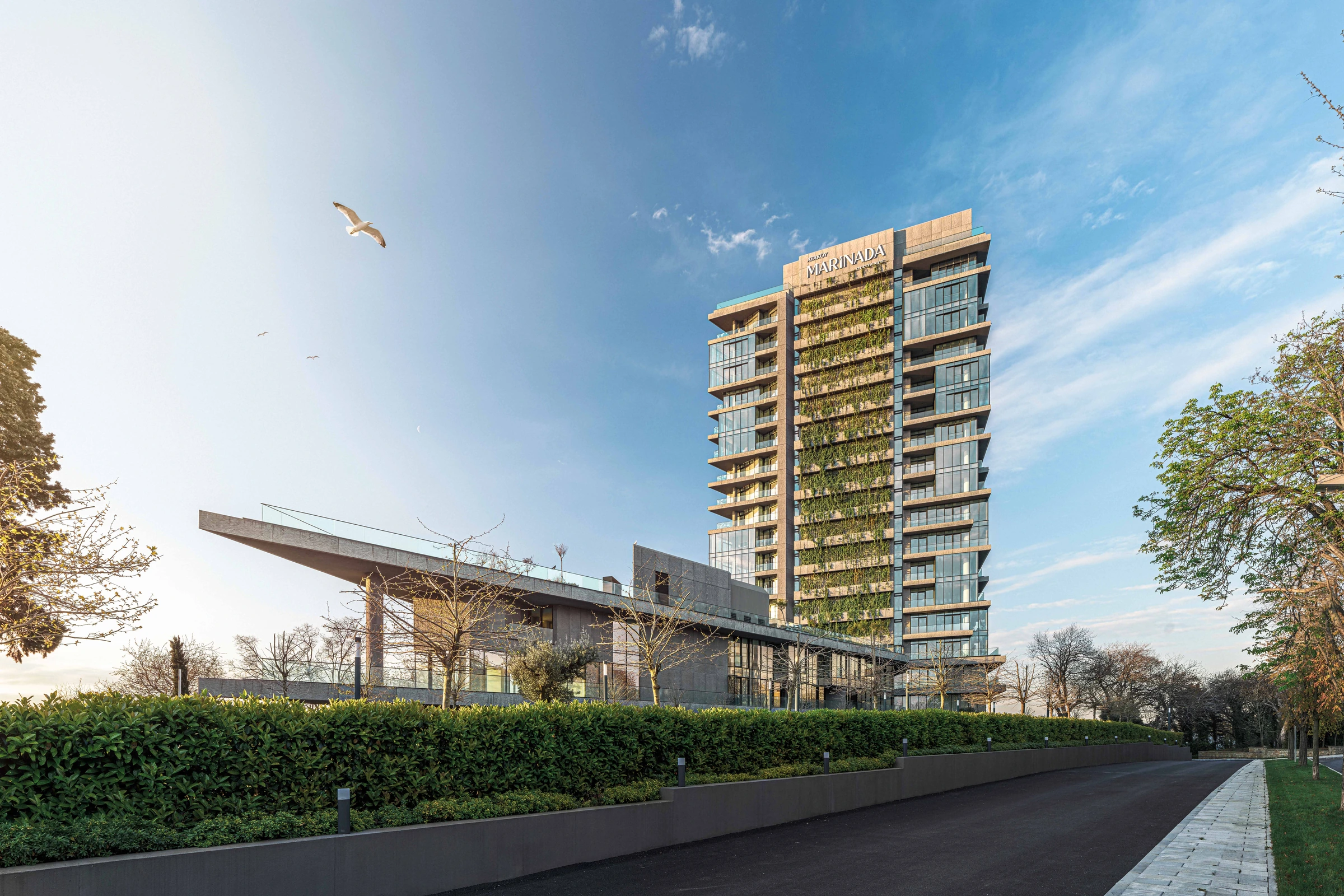 Açık mavi gökyüzü altında Marinada Residence; önünde kanat biçimli çatısıyla sosyal tesis ve yol boyunca yeşil peyzaj